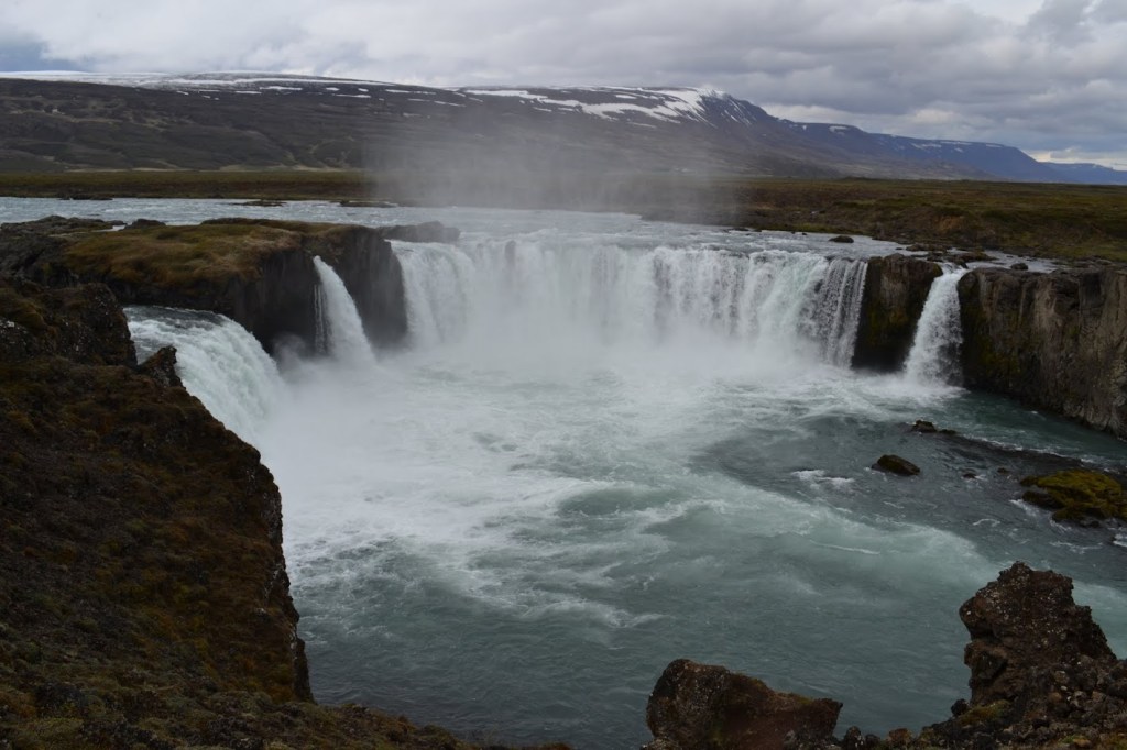 Goðafoss is a waterfall in Iceland, approximately 12 meters (39 feet) tall, but 30 meters (about 98 feet) wide. There are 3 distinct larger plunges and one smaller waterfall to the right. In this view, the snow-covered mountains are visible in the distance.