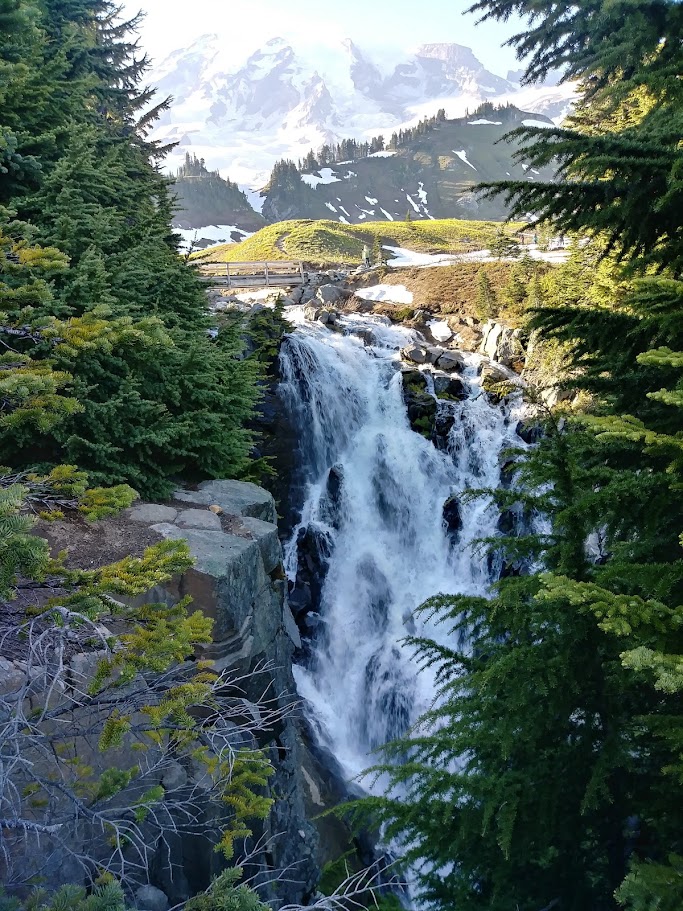 This is Myrtle Falls, a waterfall in Washington state inside Mount Rainier National Park. The waterfall is 72 feet tall. There is a bridge directly above the waterfall, and then glaciers and the top of Mount Rainier are visible. 