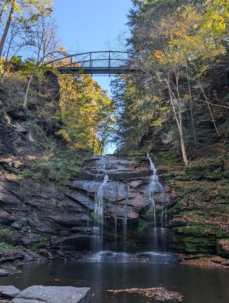 This is Rexford Falls, a 30-foot / 10 meter tall waterfall in New York. There is a bridge above the waterfall and there are trees changing colors on the rock walls in the gorge.