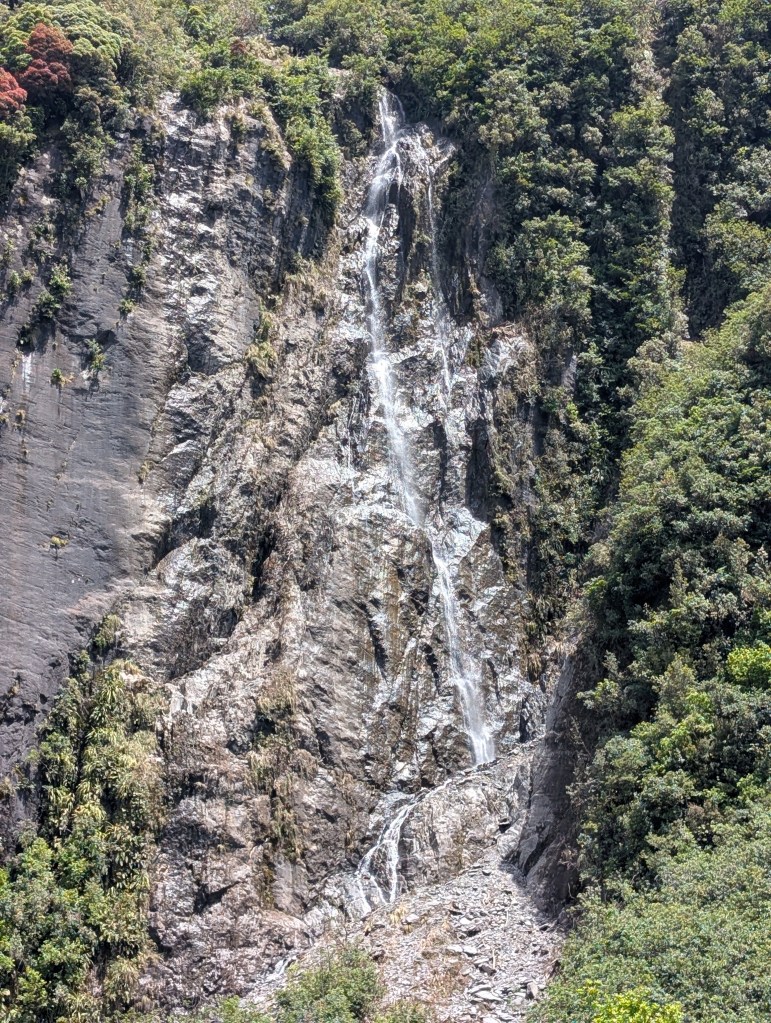 This waterfall might be Trident Creek Falls. It is a 100 foot / 30 meter tall waterfall that cascades down a rock face surrounded by trees.